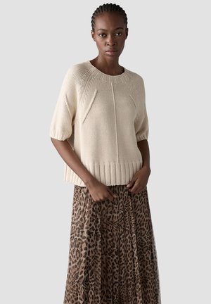 Frau trägt einen beigen kurzärmeligen Strickpullover und einen langen Rock mit Leopardenmuster, steht vor einem schlichten Hintergrund.