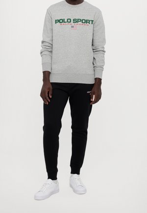 Mann trägt grauen Sweatshirt mit der Aufschrift "POLO SPORT RALPH LAUREN" und kleiner US-Flagge, schwarze Jogginghose und weiße Sneaker, steht vor weißem Hintergrund.