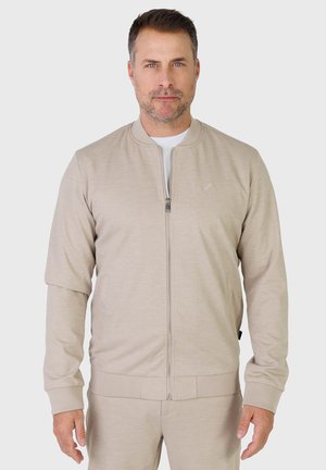 Homme aux cheveux bruns courts et à la barbe légère portant une veste beige à fermeture éclair et un pantalon assorti, debout devant un fond clair uni.