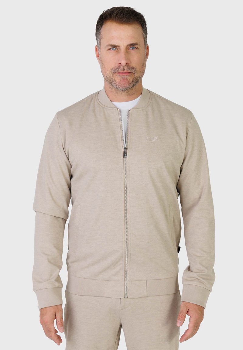 Homme aux cheveux bruns courts et à la barbe légère portant une veste beige à fermeture éclair et un pantalon assorti, debout devant un fond clair uni.