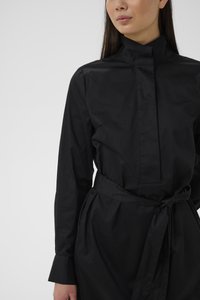 Nő hosszú ujjú fekete ruhában, magas nyakkal és megkötött derékövvel, egyszínű világos háttér előtt állva.