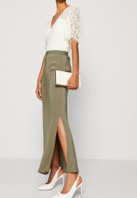 Lång grön kjol med sidoslits, vit blus i spets med korta puffärmar och djup V-ringning, kombinerat med vita högklackade skor med spetsiga tår och en clutch.