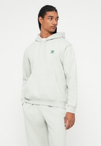Fiatal személy fonott hajjal, világosszürke Adidas kapucnis pulóvert és hozzá illő nadrágot viselve, oldalra nézve, egy sima fehér háttér előtt.