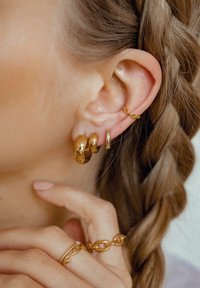 Goldene Ohrreifen in verschiedenen Größen, die eine glatte, reflektierende Oberfläche zeigen; zusätzliches goldfarbenes Kettenohrring-Accent; geflochtenes Haar sichtbar.