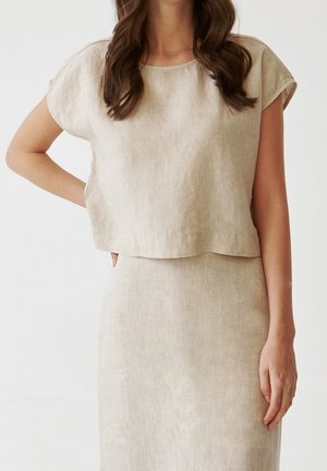 Woman wearing a beige linen short-sleeve top and matching high-waist skirt, standing with one hand on her hip.