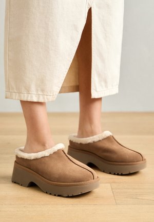 Zuecos de gamuza marrón con un forro de color crema esponjoso, puntera redondeada y suela de goma elevada. Presentan costuras visibles en la parte superior.
