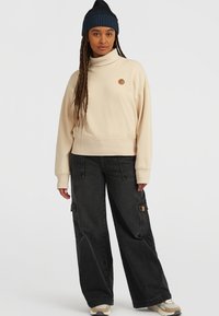 Beige sweatshirt med hög krage och logotyppatch, kombinerad med löst sittande svarta cargo byxor med fickor. Slät tyg och avspänt snitt.
