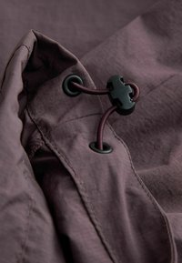 Material ușor, de culoare violet închis, cu o suprafață texturată, dotat cu un cordon de strângere cu accesorii din plastic negru și orificii pentru ajustare.