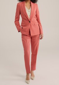 Žena v korálovém přizpůsobeném sakě a odpovídajících kalhotách, v kombinaci s svetříkem světle béžové barvy a béžovými lodičkami s špičatou špičkou a detaily na přezku.