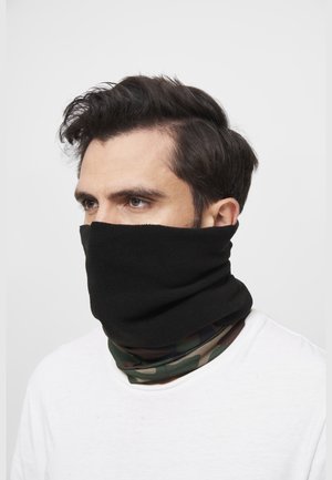 Hombre con cabello oscuro que lleva un calentador de cuello negro de forro polar sobre un pasamontañas de camuflaje y una camisa blanca, mirando hacia la izquierda.