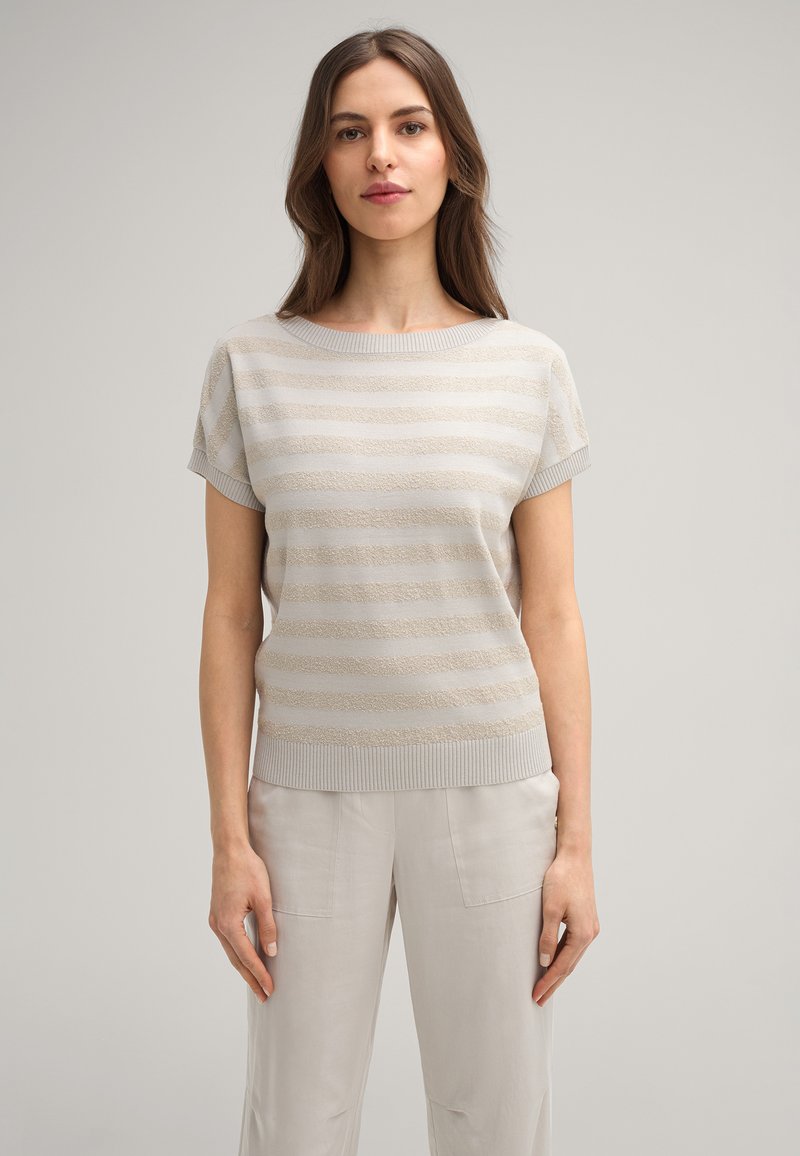 Hellbeiges Strickoberteil mit kurzen Ärmeln, das über strukturierte horizontale Streifen sowie einen gerippten Ausschnitt und Saum verfügt. Kombiniert mit hellen Hosen.