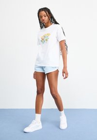 Woman with long braids wearing white "Santa Cruz" floral print T-shirt, light blue sports shorts, white socks, and white sneakers standing on blue floor.