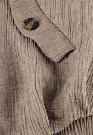 Beige ribbestrikket stoff med en mansjett som har en stor brun knapp sydd på et teksturert stoff.