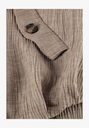 Beige ribbestrikket stoff med en mansjett som har en stor brun knapp sydd på et teksturert stoff.