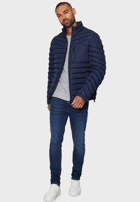 Marineblå puffjakke med quiltet tekstur, høj krave og lynlåslomme på brystet, parret med en grå skjorte og blå jeans, båret med hvide sneakers.