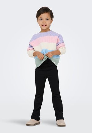 Regenbooggestreepte, geribbelde trui van gebreide stof in pasteltinten; zwarte nauwsluitende broek; beige schoenen; kind houdt de zoom van de trui vast.