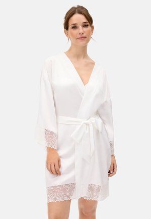 Mujer vistiendo una bata cruzada de satén blanca con borde de encaje en las mangas y el dobladillo, atada a la cintura con un lazo, de pie contra un fondo claro.