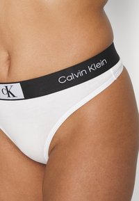 White cotton bikini underwear with a black waistband featuring "Calvin Klein" logo in white. Smooth texture, fitted shape.
