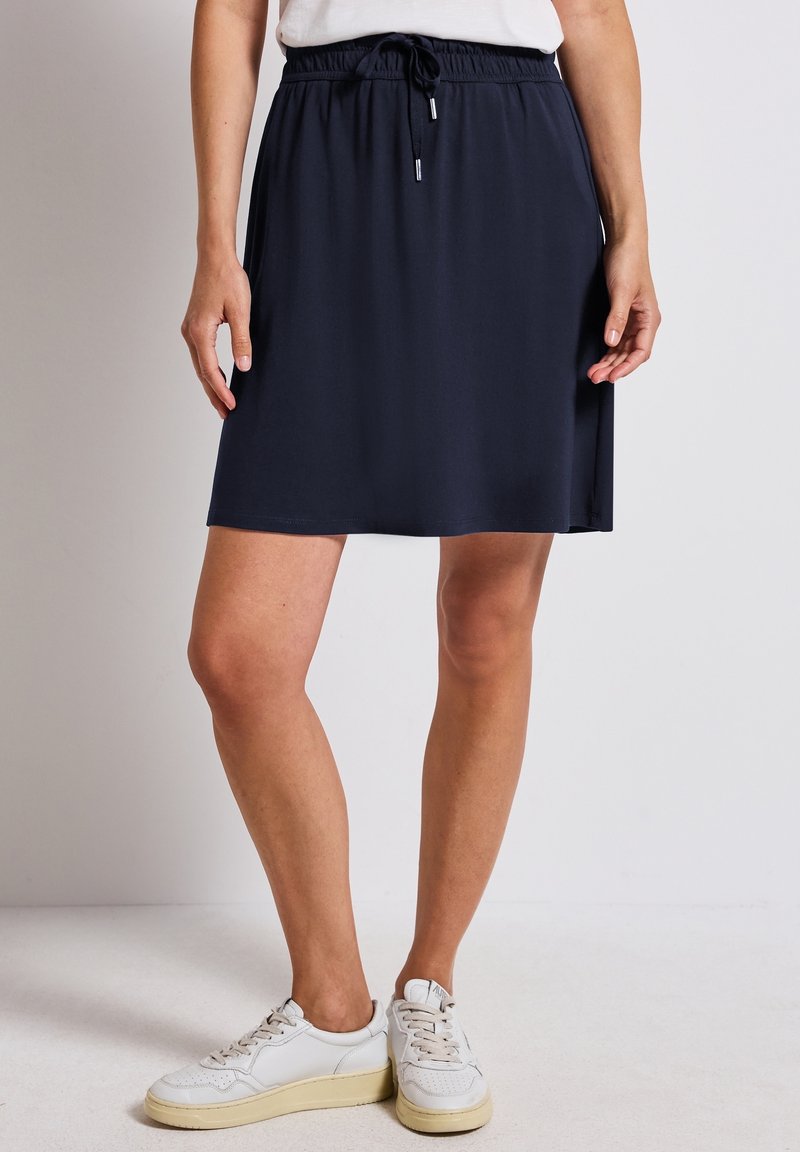 Femme portant une jupe bleu marine mi-longue avec une taille élastique et un cordon de serrage, associée à des baskets blanches et un haut blanc.
