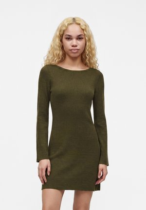 Jeune femme aux longs cheveux blonds bouclés portant une robe en maille côtelée moulante à manches longues de couleur vert olive, debout devant un fond uni.