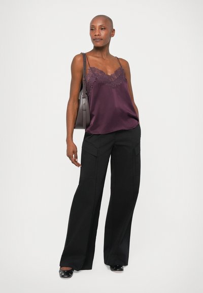 Satiinlilla kampsun, millel on pitsäärised, koos laia säärtega mustade pükstega, millel on kottide taskud. Mudel hoiab halli õlakotti.