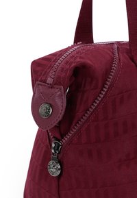 Burgundy shoulder bag with a textured fabric, featuring a zippered closure, circular leather accent with embossed logo, and sturdy straps.