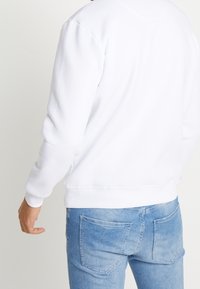 Hombre vistiendo un sudadera blanca sencilla y jeans de mezclilla azul claros, visto desde atrás con un brazo ligeramente doblado.