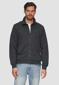 Chaqueta negra con cremallera en tejido suave, con un collar alto, puños y dobladillo acanalados, con una textura lisa y un detalle de logo minimalista.