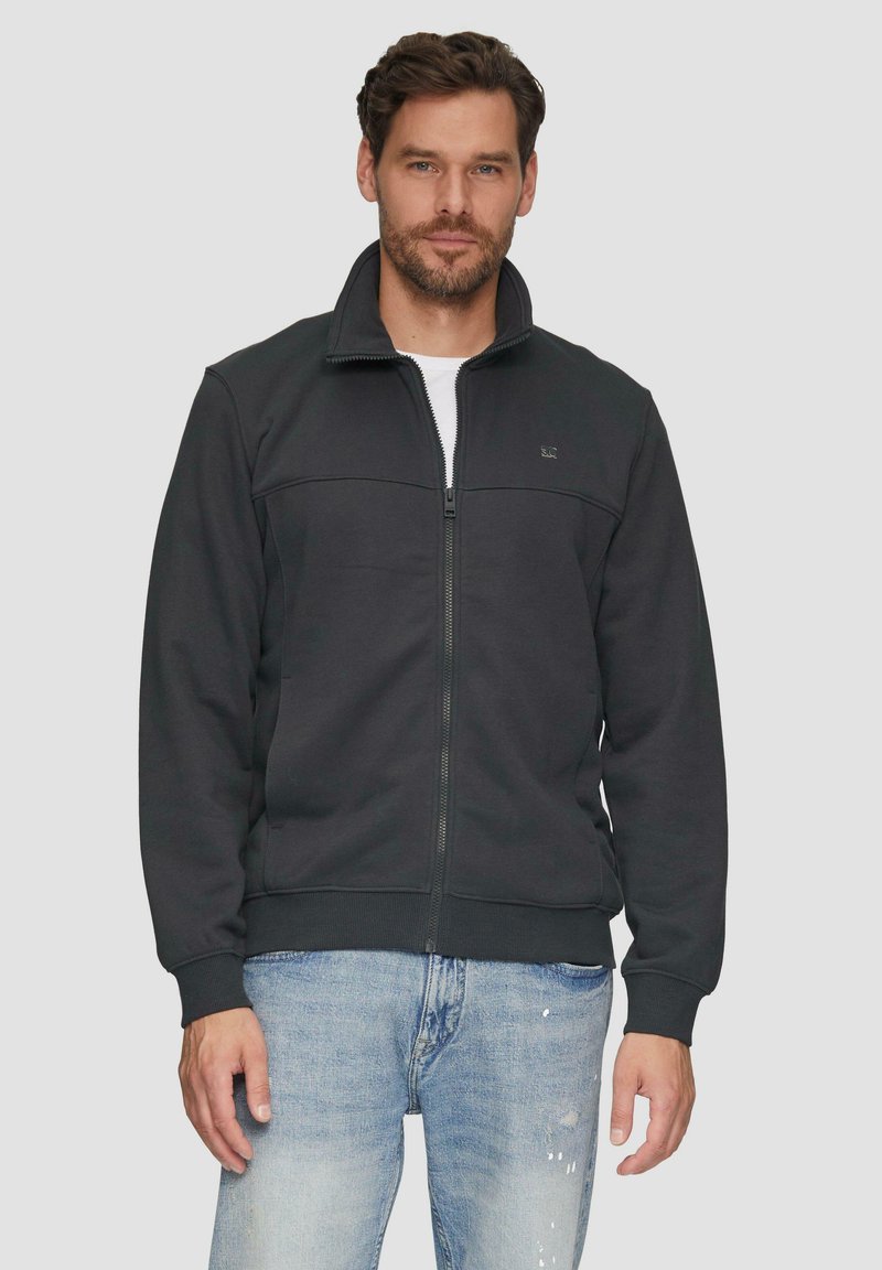 Chaqueta negra con cremallera en tejido suave, con un collar alto, puños y dobladillo acanalados, con una textura lisa y un detalle de logo minimalista.