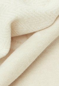 Beige gebreide stof met een zachte textuur en subtiele schaduwvariaties, met plooien die een gelaagd uiterlijk vormen.