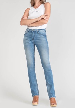 Kobieta w jasnoniebieskich obcisłych dżinsach, białym bezrękawnym topie z napisem oraz beżowych sandałach na platformie, stojąca z założonymi ramionami.