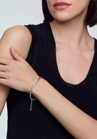 Srebrna bransoletka o fakturowanym, falistym wzorze, noszona na nagim nadgarstku. Osoba nosząca ma na sobie czarny, dziany top bez rękawów z okrągłym dekoltem.