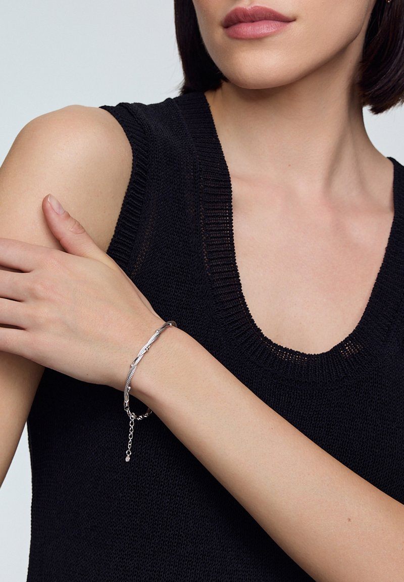 Srebrna bransoletka o fakturowanym, falistym wzorze, noszona na nagim nadgarstku. Osoba nosząca ma na sobie czarny, dziany top bez rękawów z okrągłym dekoltem.