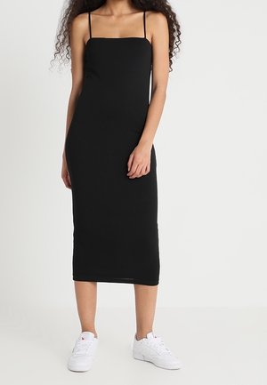 Femme portant une robe midi noire ajustée sans manches avec de fines bretelles et des baskets blanches, debout devant un fond blanc uni.