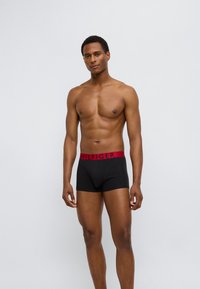 Μαύρα ανδρικά εσώρουχα τύπου boxer με κόκκινη ελαστική μέση που φέρει το λογότυπο "HILFIGER". Το υλικό φαίνεται λείο· το σχέδιο είναι εφαρμοστό με μέση μεσαίου ύψους.