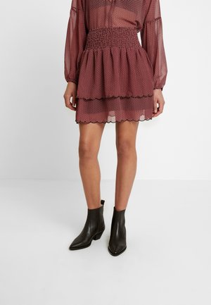 Jupe mini rouge à pois avec des volants superposés et des bords festonnés, portée avec des bottines noires pointues et une blouse translucide à manches longues.