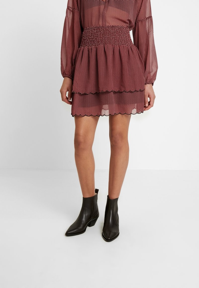 Röd prickig mini kjol med lager av volanger och scallopade kanter, bärs med svarta spetsiga ankelstövlar och en genomskinlig blus med långa ärmar.