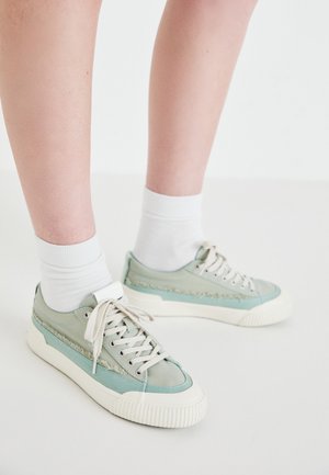 Lichtgroene sneakers met een gestructureerde grijsbruine accen, witte veters en een ribbelige ivoorkleurige zool, gedragen met witte sokken. Platte, low-top ontwerp.