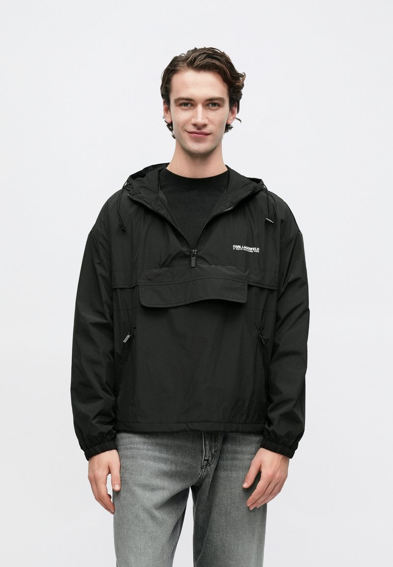 Νεαρός άνδρας που φοράει μαύρο αδιάβροχο με μισή φερμουάρ και κουκούλα, καθώς και γκρι τζιν, στέκεται μπροστά σε ένα απλό φωτεινό φόντο.