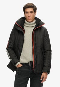 Czarna pikowana kurtka z pomarańczowym zamkiem i akcentami przy kołnierzu. Nosi się ją na jasnoszarym swetrze dzianinowym. Zawiera detal z logo na rękawie.