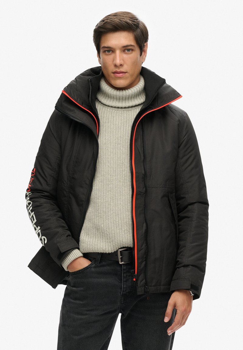 Chaqueta acolchada negra con cremallera y detalles del cuello en naranja. Lleva encima un suéter de punto gris claro. Presenta un detalle de logo en la manga.