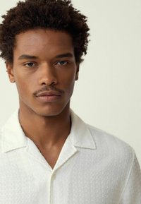 White short-sleeved shirt with a textured pattern, featuring a notched collar and button closure, worn by a person with curly hair.