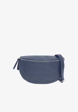 Femme souriante tenant un sac bandoulière en cuir bleu marine, portant un haut noir à manches longues et un pantalon en cuir noir, debout devant un fond blanc.