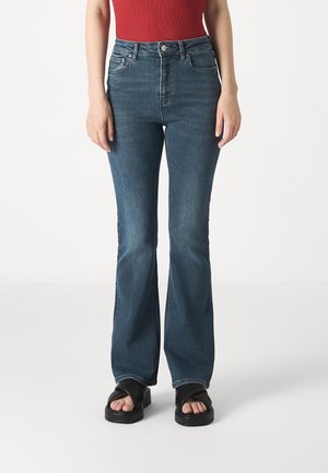 Femme portant un jean flare bleu taille haute, un top rouge sans manches et des sandales à plateforme noires à bout ouvert, debout sur un fond blanc.