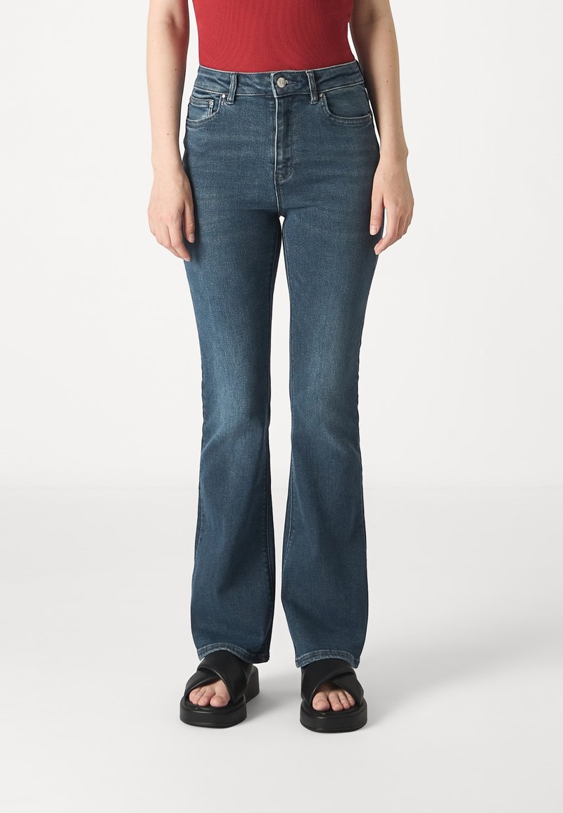 Femme portant un jean flare bleu taille haute, un top rouge sans manches et des sandales à plateforme noires à bout ouvert, debout sur un fond blanc.