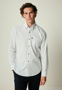 Camisa blanca de manga larga con un pequeño patrón geométrico en azul y verde. Presenta un cuello abotonado y un bolsillo en el pecho.