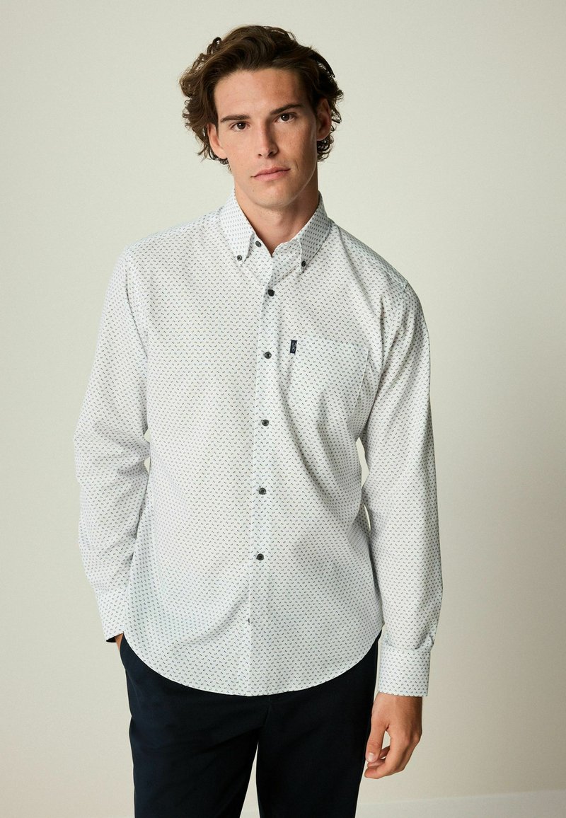 Camisa blanca de manga larga con un pequeño patrón geométrico en azul y verde. Presenta un cuello abotonado y un bolsillo en el pecho.