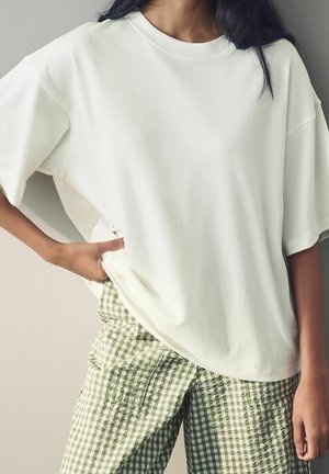 Kobieta ubrana w luźny biały T-shirt z krótkim rękawem oraz jasnozielone i białe spodnie w kratę, ręka oparta na biodrze.