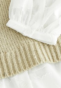 Beige strikket sweater med tekstureret stof og en hvid, gennemsigtig bluse under, med en knappet hvid manchet og delikate stof detaljer.