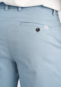 Ljust blå texturerade byxor har en liten bakficka med svart knappstängning och ett märkeslabel nära midjan.